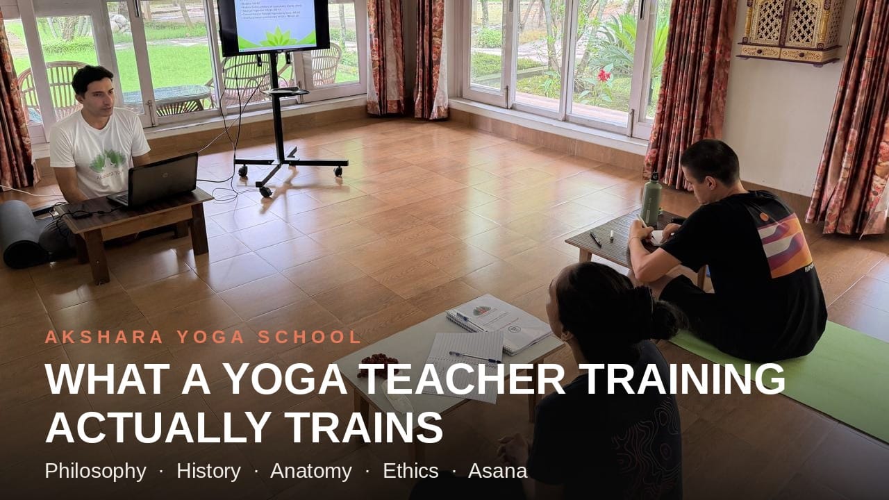 Two yoga teacher training students at Akshara Yoga School sitting on mats and low tables, taking notes during a classroom lecture with a presentation screen.